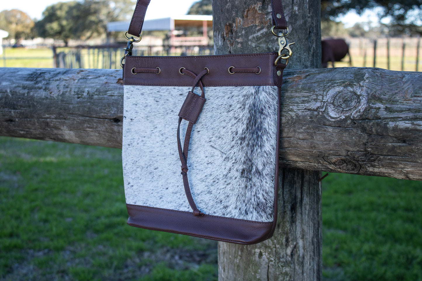The front of the purse is shown against a wooden fence post. The purse is genuine brown leather and black and gray cowhide. It has a leather drawstring located on the front. The purses dimensions are 12x12.