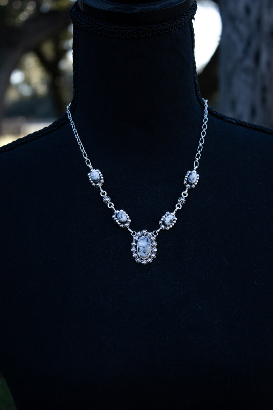 A black mannequin in an outdoor setting with a necklace sitting on it's neck. The necklace is 18" in length from one end to one end and has 5 stones of white buffalo turquoise which are white with black matrix and they are all oval shape, the biggest stone being in the center and the smaller stones on the left and right side 2 each. The necklace is set in .925 silver. There are silver dividers between the stones. Southwestern style jewelry.