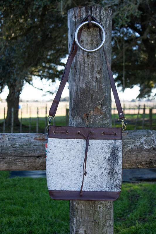 Genuine brown leather and gray and black cowhide purse, sitting a fencepost in the country on a sunny day. The purse is bucket style with a removable, adjustable shoulder strap. There is a leather draw string located in the front. The hardware on purse is gold colored.